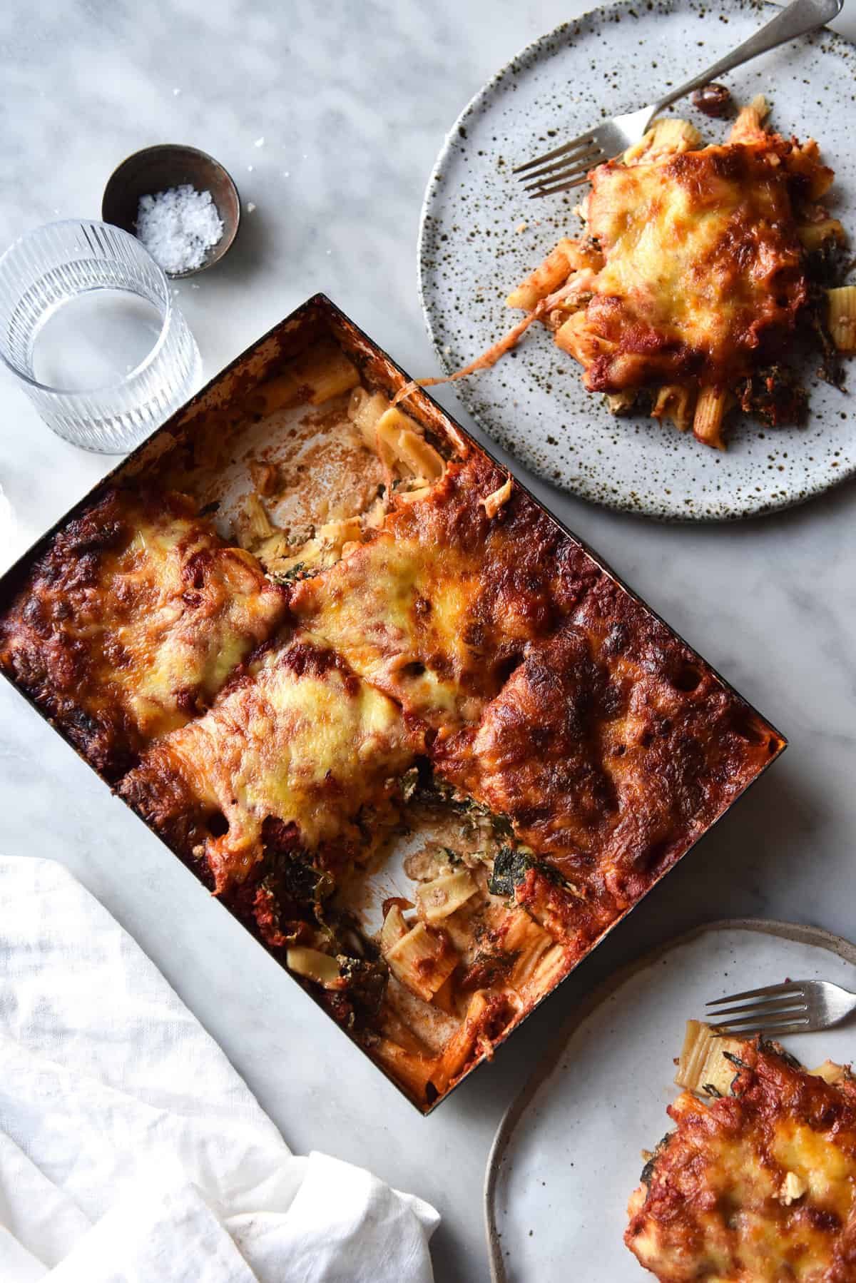 An aerial image of a gluten free pasta bake on a white marble table. The bake is surrounded by extra plates of pasta bake and linen tablecloths. 