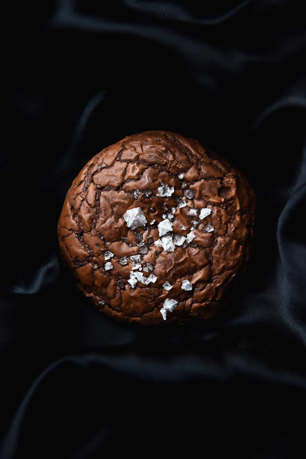 A dark aerial image of a crackled gluten free brownie cookie topped with sea salt flakes. The cookie sits atop a black silky backdrop.