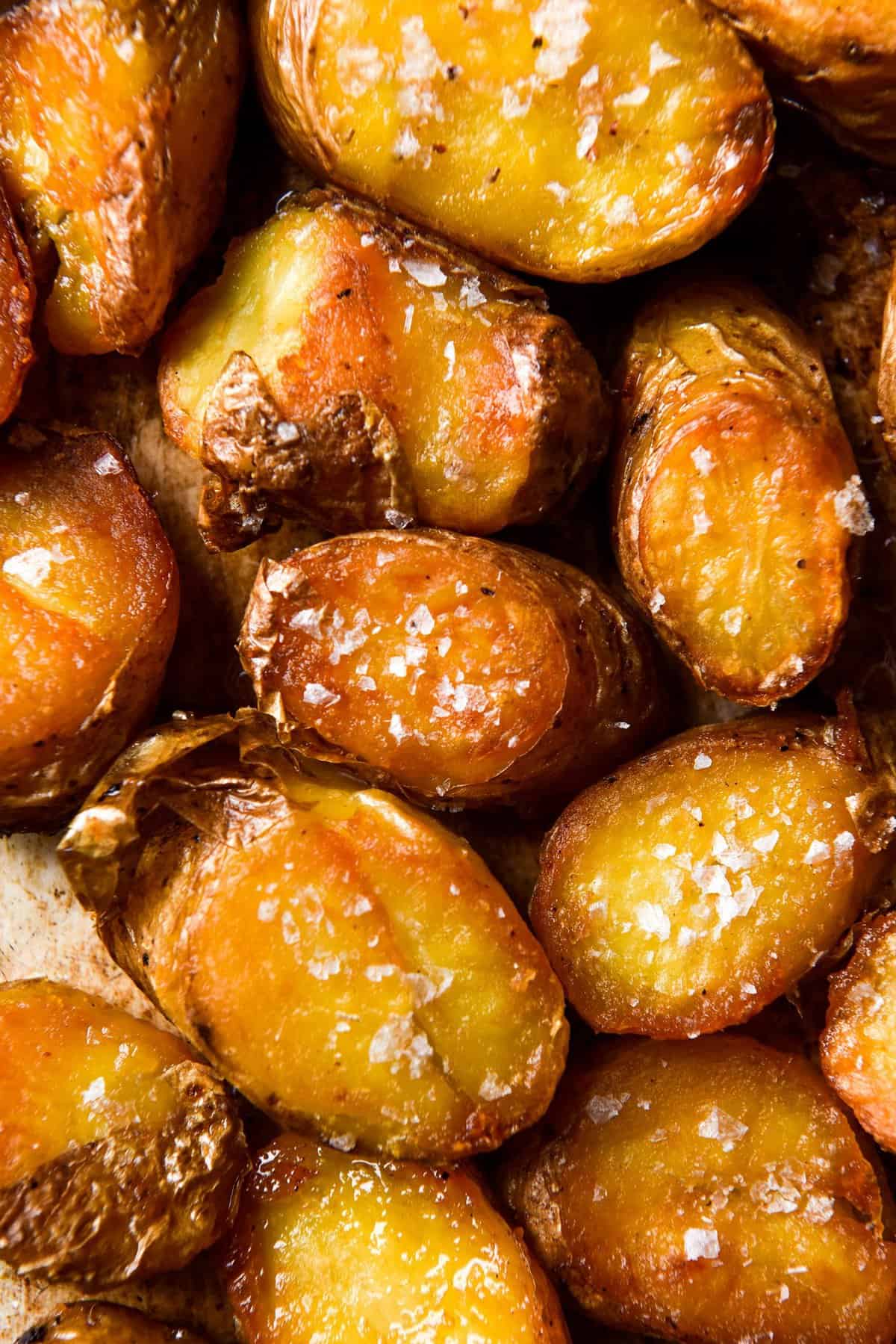 An aerial macro close up image of crispy roasted potatoes on a rusty baking tray. The potatoes are golden and topped with sea salt flakes. 