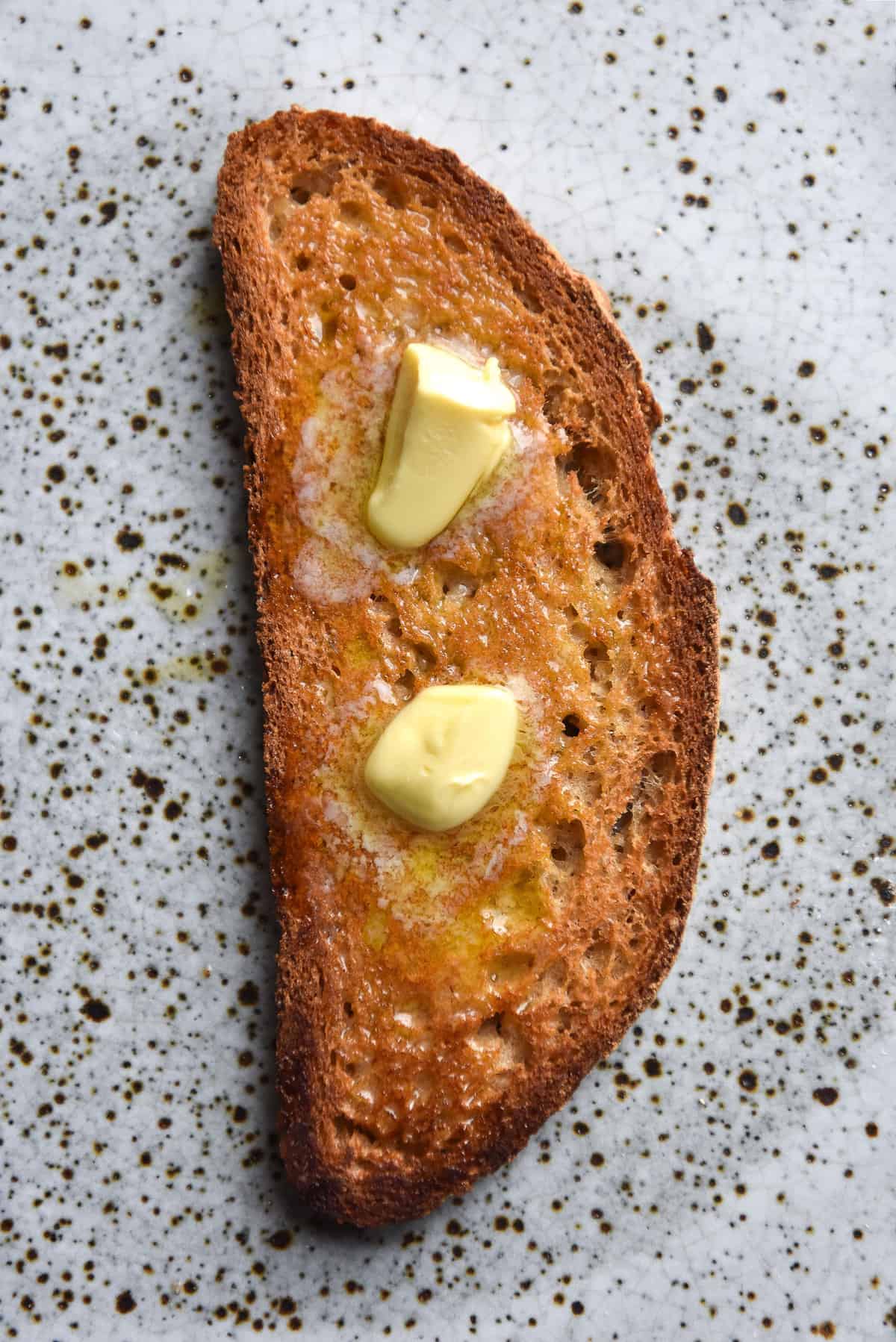 An aerial image of a slice of gluten free sourdough toast on a white speckled ceramic plate. The toast is golden brown and topped with melting butter.