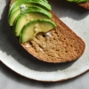 An aerial image of gluten free sourdough bread toasted and topped with sliced avocado. The toast sits on a white ceramic plate atop a white marble table.