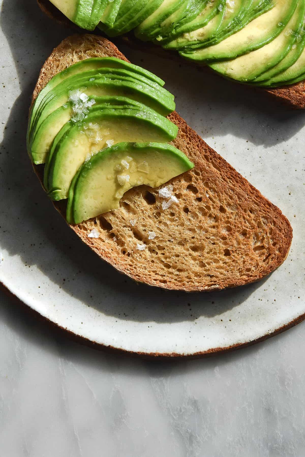 An aerial image of gluten free sourdough bread toasted and topped with sliced avocado. The toast sits on a white ceramic plate atop a white marble table.