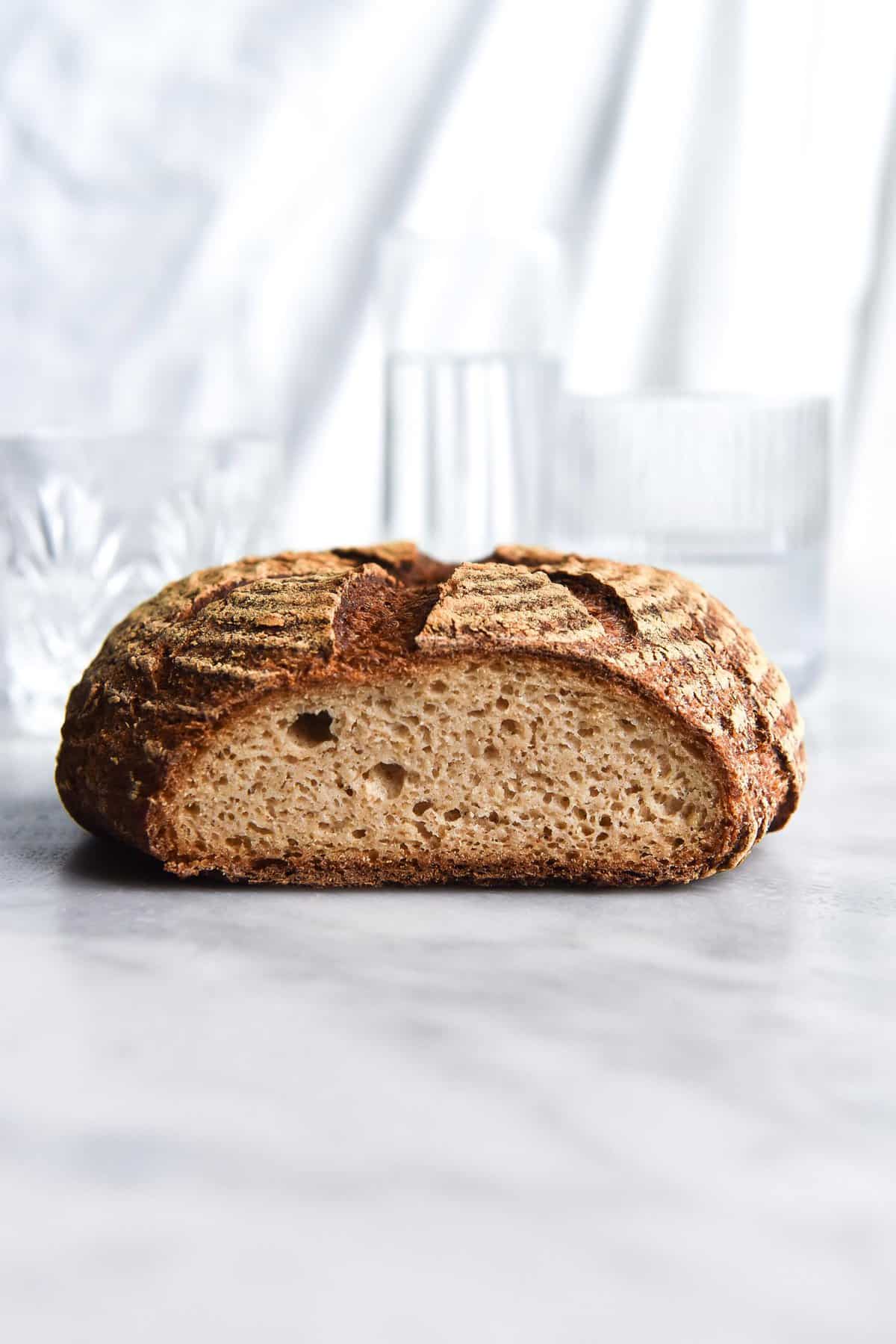 A side on view of a loaf of gluten free sourdough bread that has been sliced to reveal the crumb. It sits on a white marble table against a white backdrop and glasses of water sit in the background. 