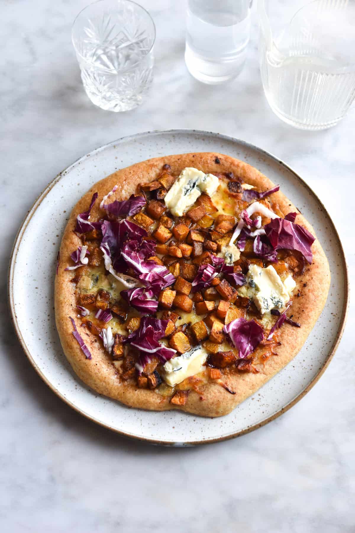 An aerial image of a gluten free pizza topped with roasted pumpkin, blue cheese and radicchio. The pizza sits atop a white ceramic plate on a white marble table. 