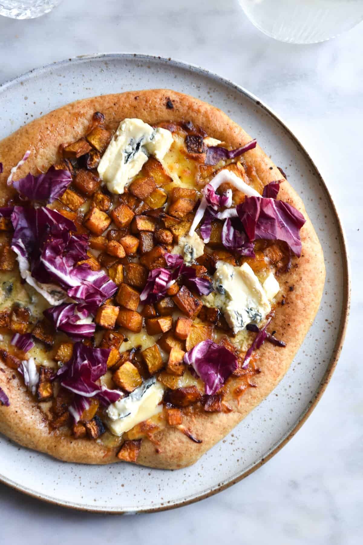 An aerial view of a gluten free pizza base on a white ceramic plate. The pizza is topped with roasted pumpkin, radicchio, blue cheese and a white pizza base.