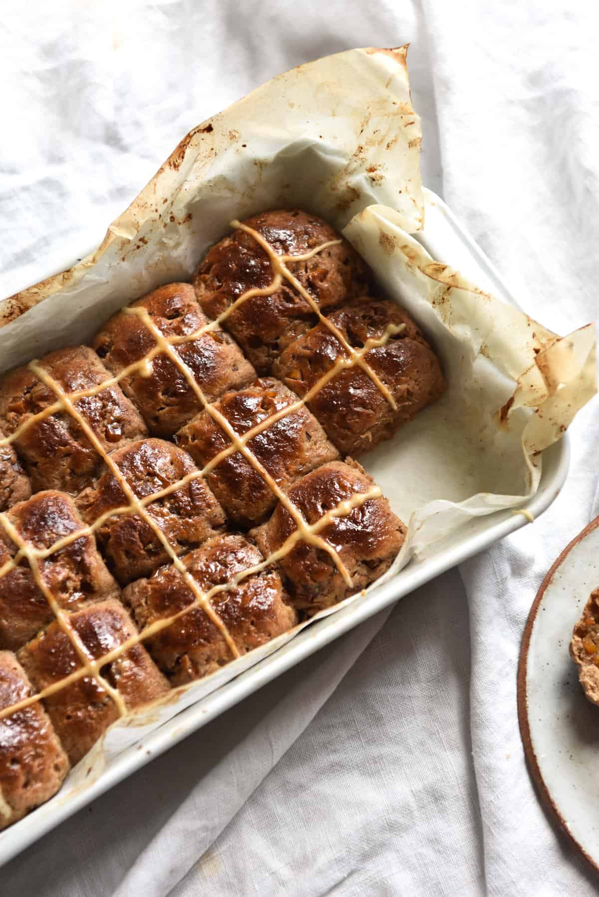 Gluten free sourdough hot cross buns linen tablecloth. One bun is removed and sits on a white ceramic plate to the bottom right of the baking dish. 