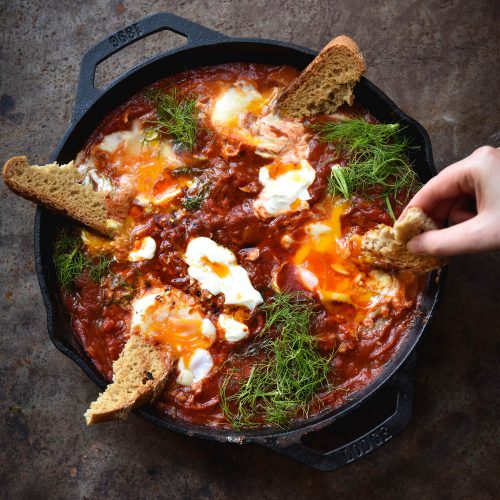 FODMAP friendly shakshuka with roasted red capsicum and haloumi in a black skillet on a rusty red backdrop. A hand extends from the right side to dip gluten free sourdough into the jammy eggs