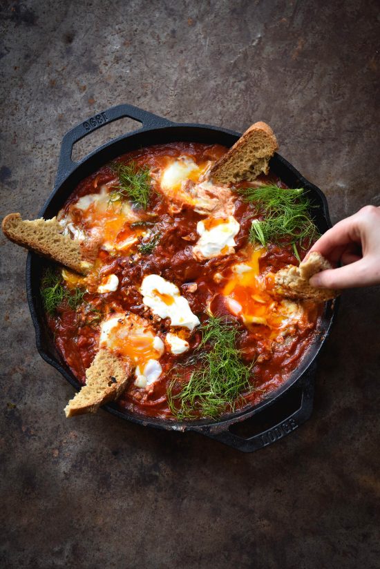 FODMAP friendly shakshuka with roasted red capsicum and haloumi in a black skillet on a rusty red backdrop. A hand extends from the right side to dip gluten free sourdough into the jammy eggs