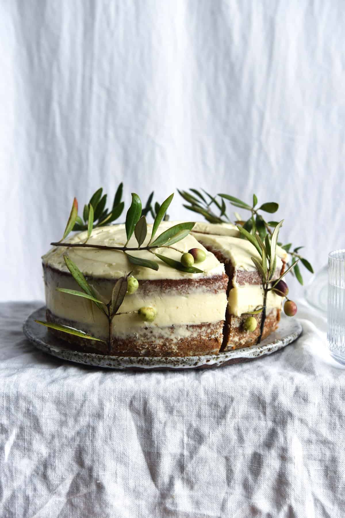 A side on view of a gluten free carrot cake. The cake is a layer cake, topped with naked cream cheese icing. The sides of the cake are decorated with olive branches. The cake sits atop a white speckled ceramic plate on a white linen backdrop