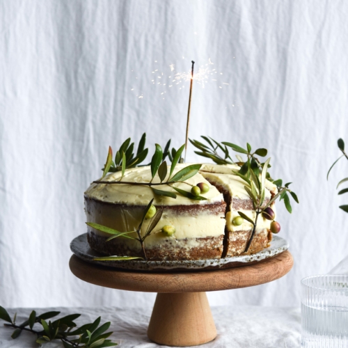 A side on view of a gluten free carrot cake. The cake is a layer cake, topped with naked cream cheese icing. The sides of the cake are decorated with olive branches, and a lit sparkler stands in the middle of the cake. The cake sits on a wooden cake stand on a white linen backdrop