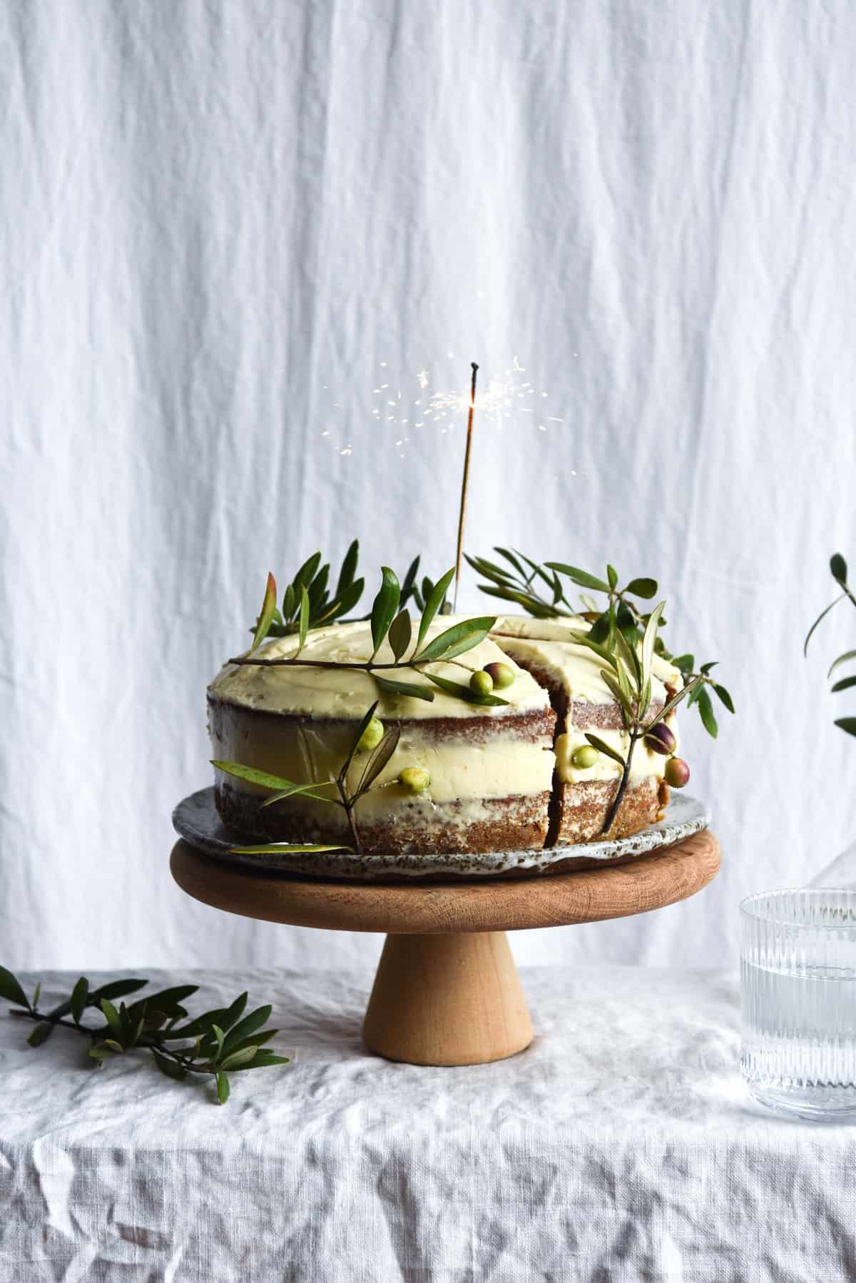 A side on view of a gluten free carrot cake. The cake is a layer cake, topped with naked cream cheese icing. The sides of the cake are decorated with olive branches, and a lit sparkler stands in the middle of the cake. The cake sits on a wooden cake stand on a white linen backdrop