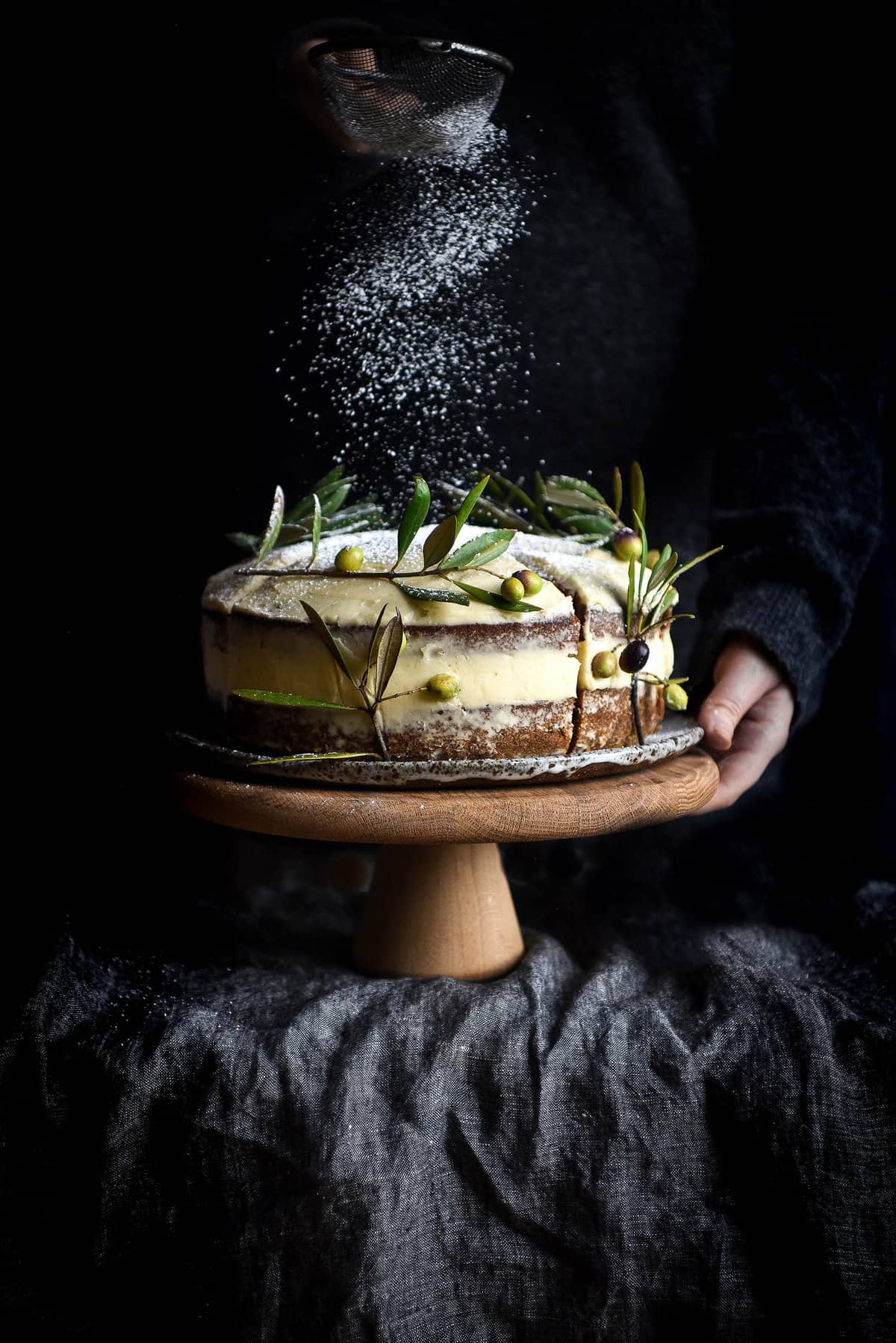 A side on view of a gluten free carrot cake on a wooden stand against a dark backdrop. A person stands behind the cake and sprinkles icing down onto the cake. The cake is covered in lactose free cream cheese icing and decorated with olive branches