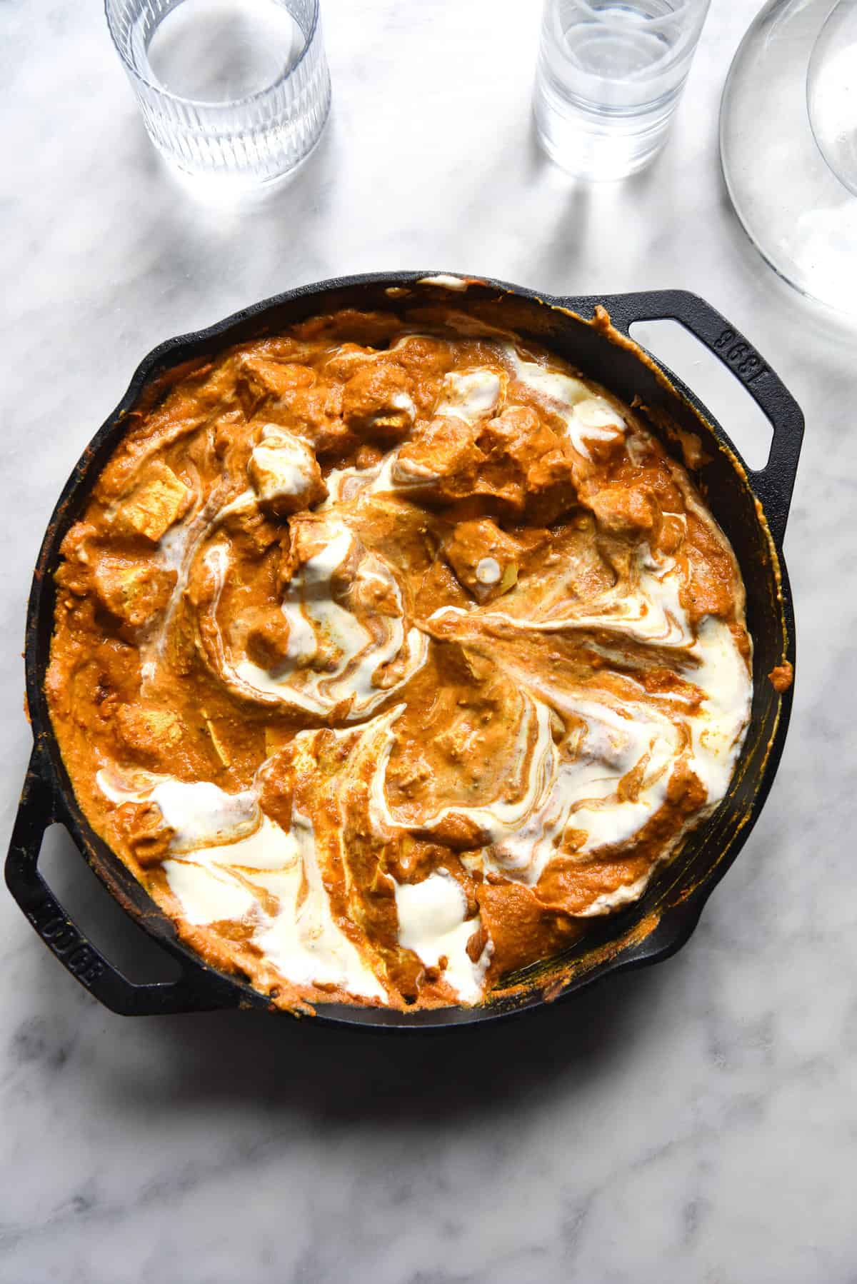 An aerial view of a skillet filled with FODMAP friendly vegetarian curry. The vibrant orange curry gravy is swirled with white coconut milk, creating a beautiful marbled effect. Pieces of tofu poke out from the gravy. The skillet is set on a white marble backdrop, and some glasses of water sit in the top of the image.