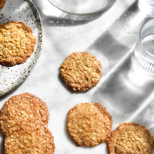 An aerial view of a white marble sunlit table topped with gluten free ANZAC biscuits and glasses of water