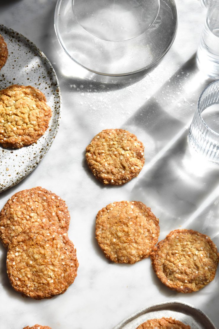 An aerial view of a white marble sunlit table topped with gluten free ANZAC biscuits and glasses of water