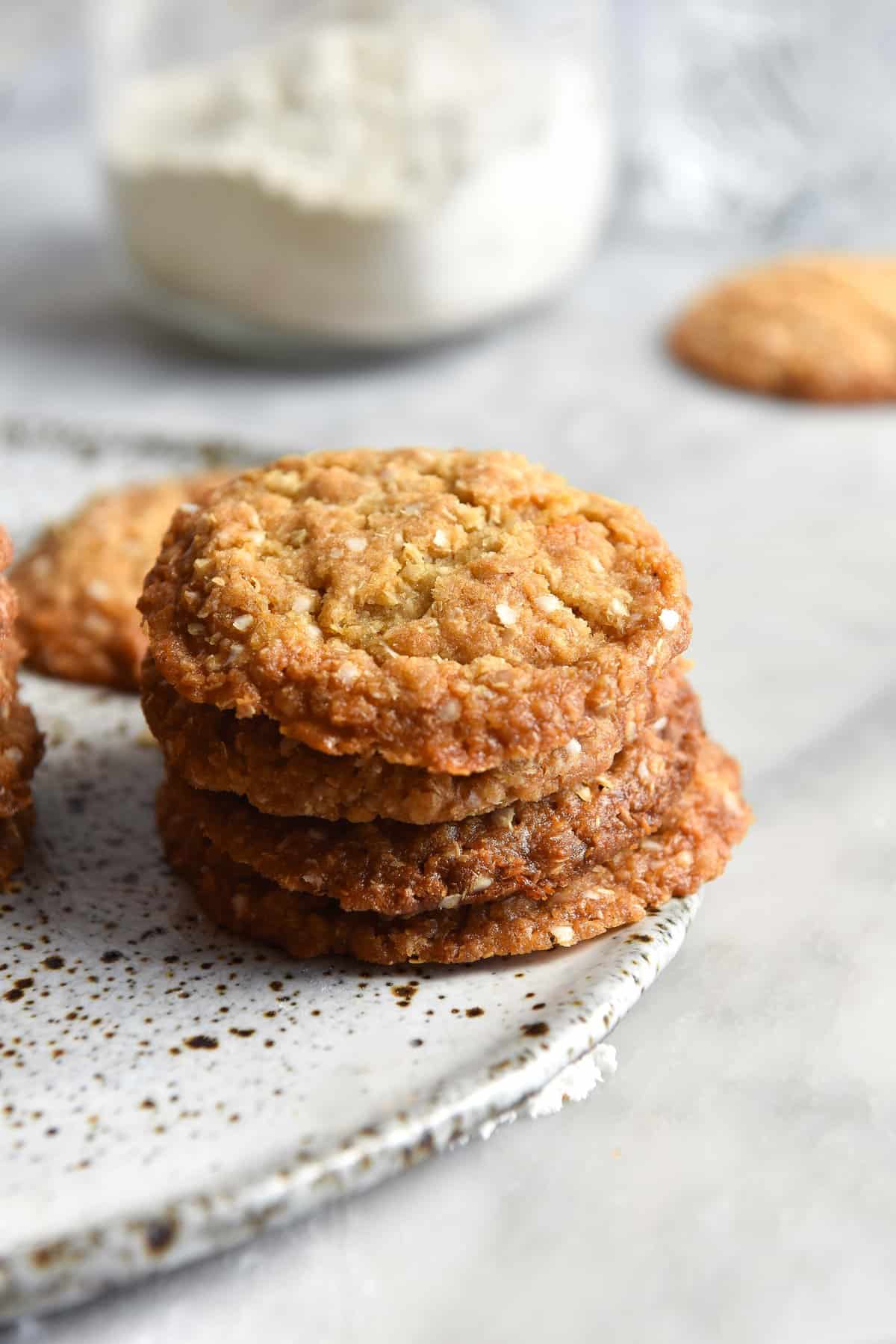 A side on image of gluten free ANZAC biscuits on a white speckled ceramic plate atop a white marble table. 