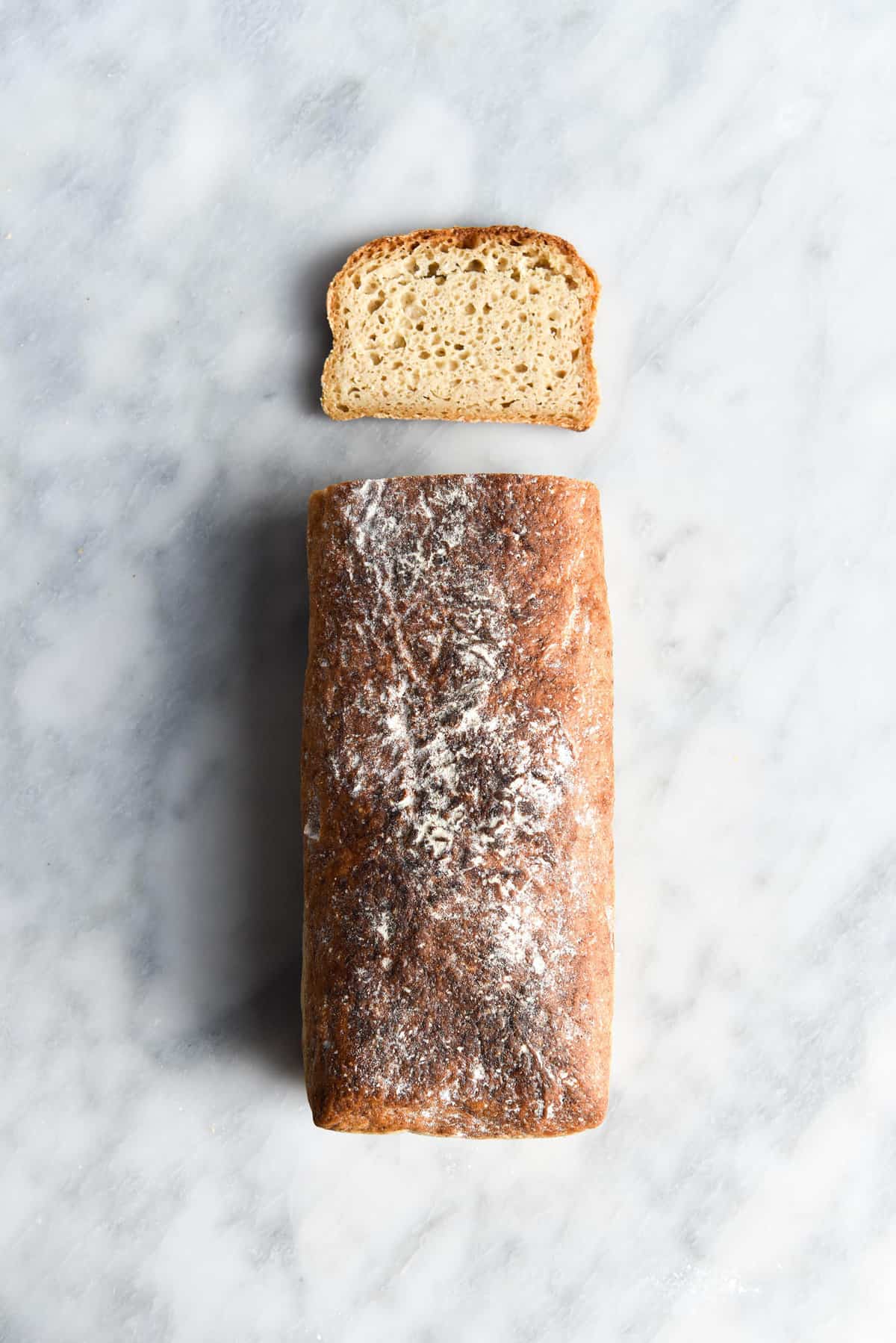 An aerial image of a loaf of lightly floured gluten free sourdough bread atop a white marble table.