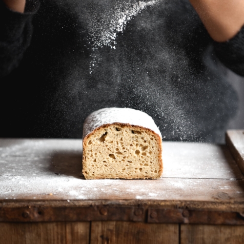 A side on view of a loaf of gluten free white bread on a wooden table. A person in a dark grey jumper stands behind the loaf and sprinkles flour down onto the bread.