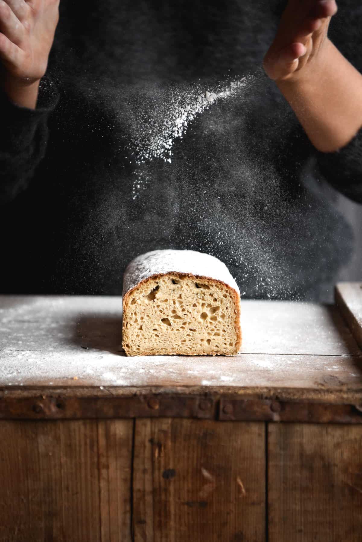 A side on view of a loaf of gluten free white bread on a wooden table. A person in a dark grey jumper stands behind the loaf and sprinkles flour down onto the bread.