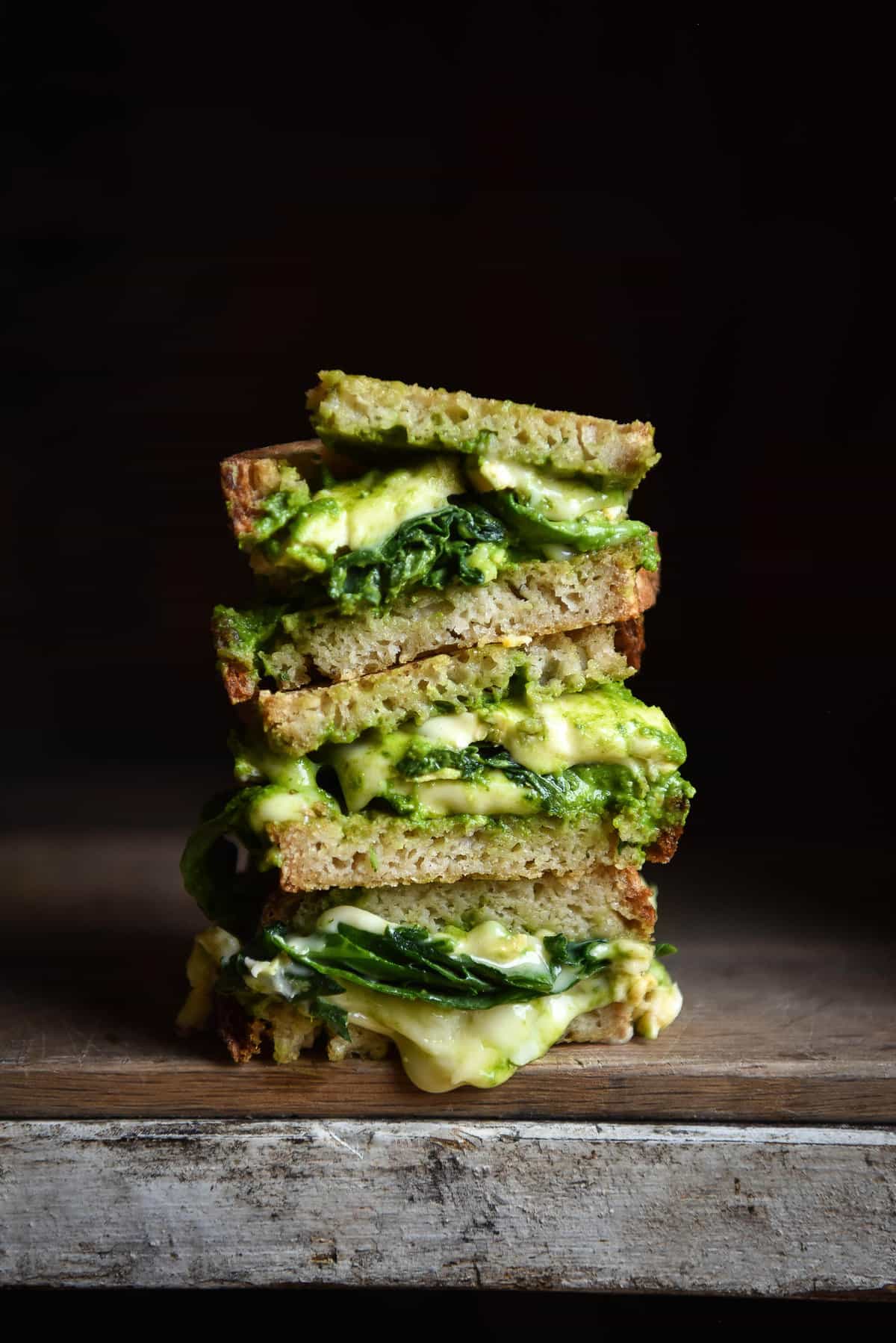 A moody side on image of a stack of sliced gluten free sourdough toasties with zingy rocket pesto and melty cheese. 