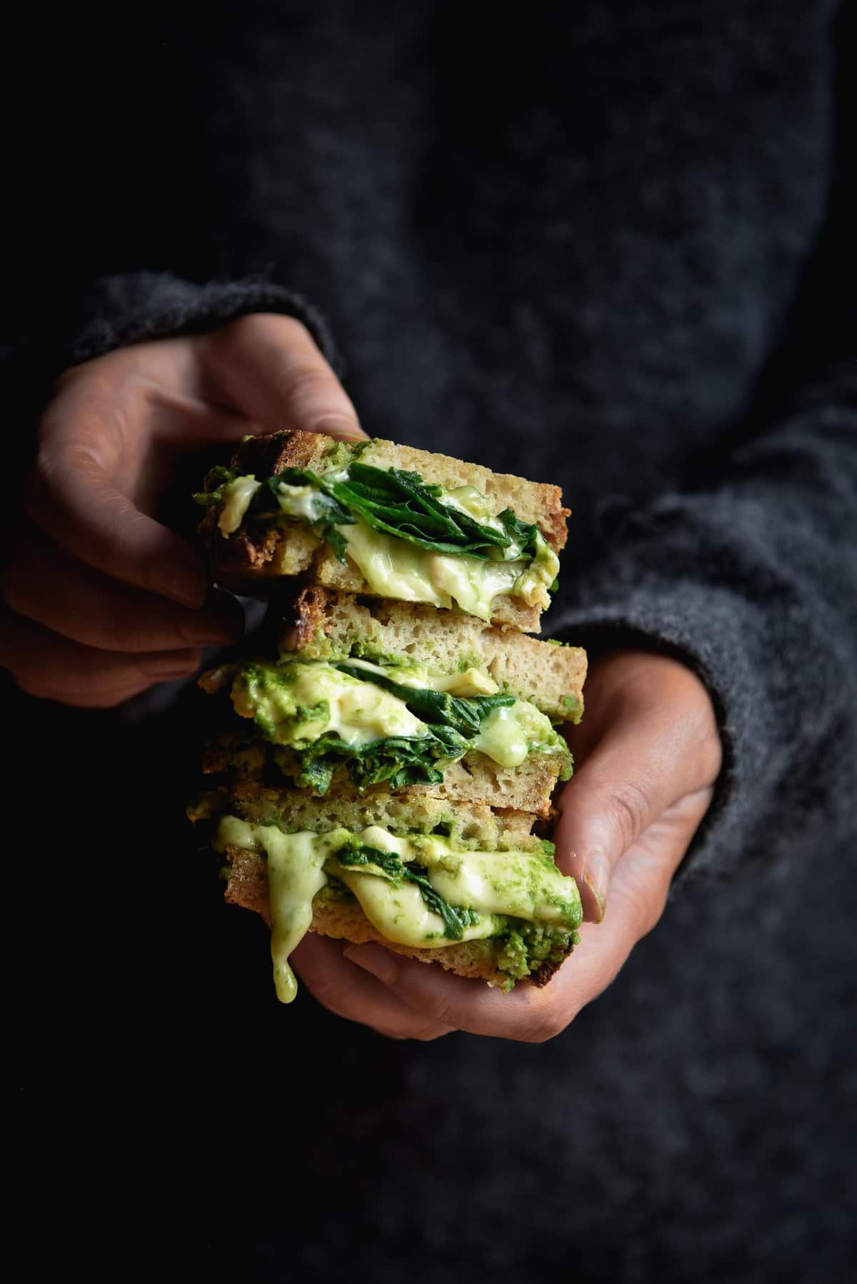 A side on moody photo of a person in a dark grey fluffy jumper holding three slices of a green pesto cheese toastie in a stack. The melty cheese oozes out of the stack and mingles with the pesto