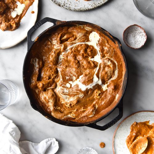 An aerial view of a skillet filled with FODMAP friendly curry that has been topped with a swirl of cream. The skillet sits atop a white marble table which is surrounded by plates of curry, water glasses and a white linen tablecloth.