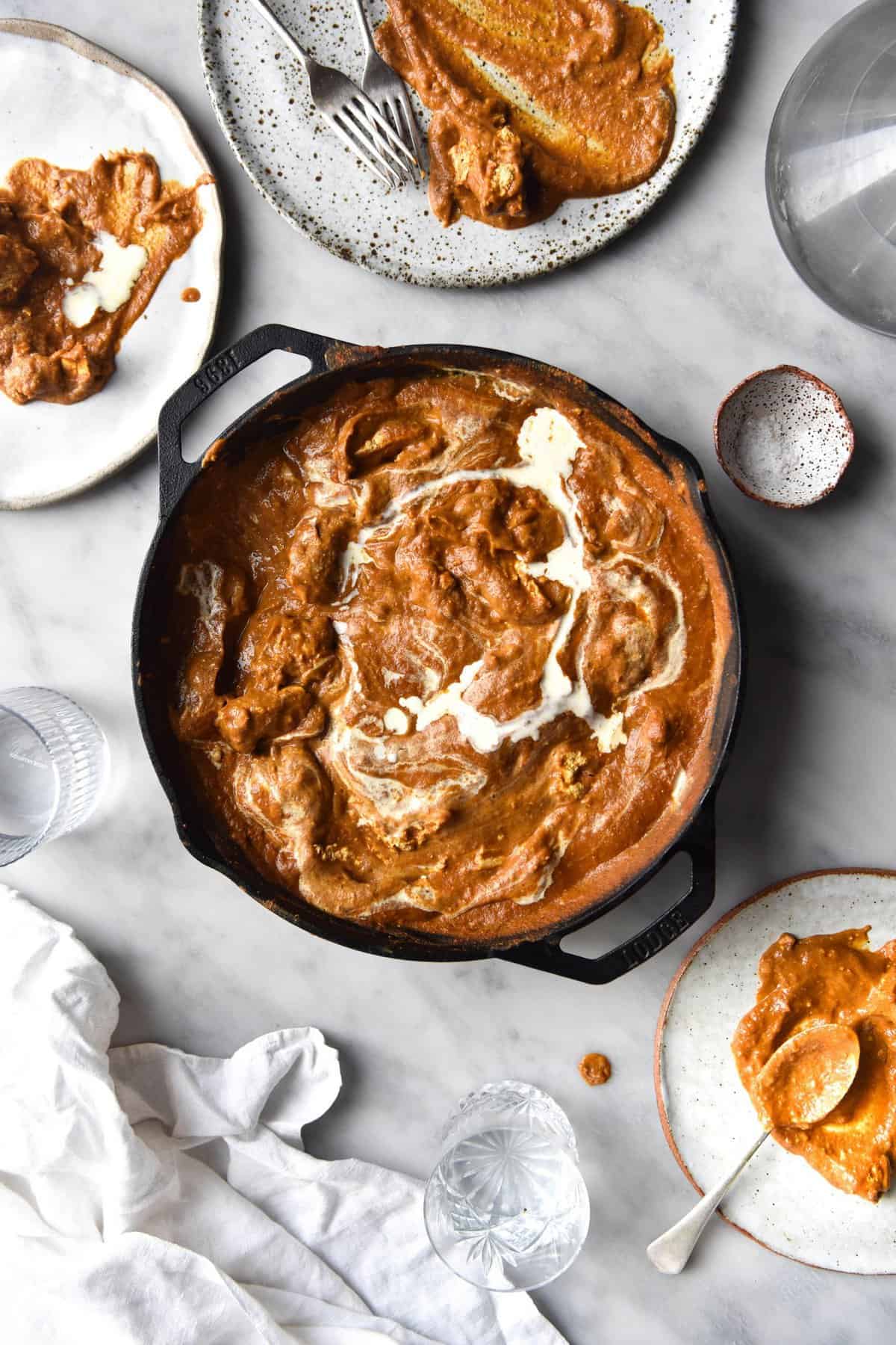 An aerial view of a skillet filled with FODMAP friendly curry that has been topped with a swirl of cream. The skillet sits atop a white marble table which is surrounded by plates of curry, water glasses and a white linen tablecloth.