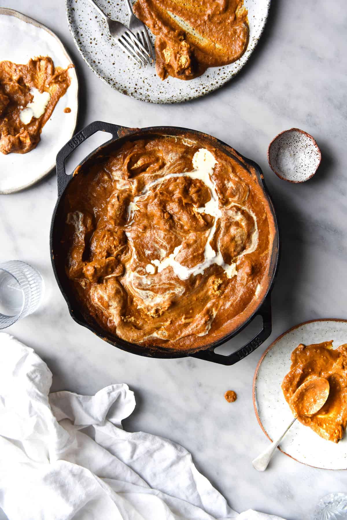 An aerial view of a skillet filled with FODMAP friendly curry that has been topped with a swirl of cream. The skillet sits atop a white marble table which is surrounded by plates of curry, water glasses and a white linen tablecloth.