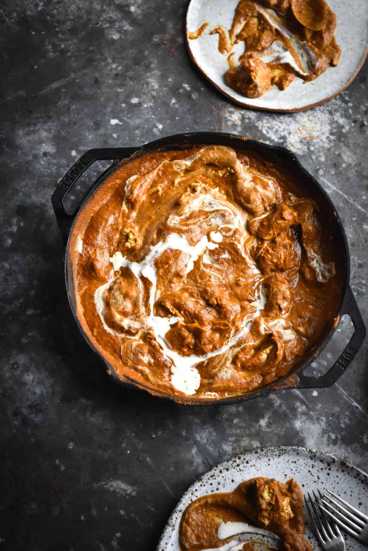 An aerial image of a skillet filled with FODMAP friendly paneer curry. The curry is a deep orange and is swirled with cream. It sits atop a medium blue steel mottled backdrop, surrounded by water glasses and extra plates
