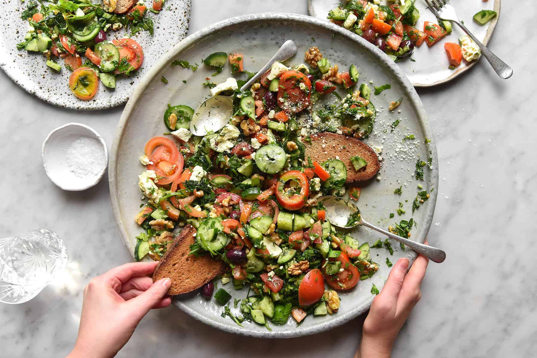 An aerial view of a cucumber and tomato salad atop a white marble table. Two smaller plates sit to the top left and top right of the main salad plate, and two hands extend over the salad to dip a piece of bread into the dressing.