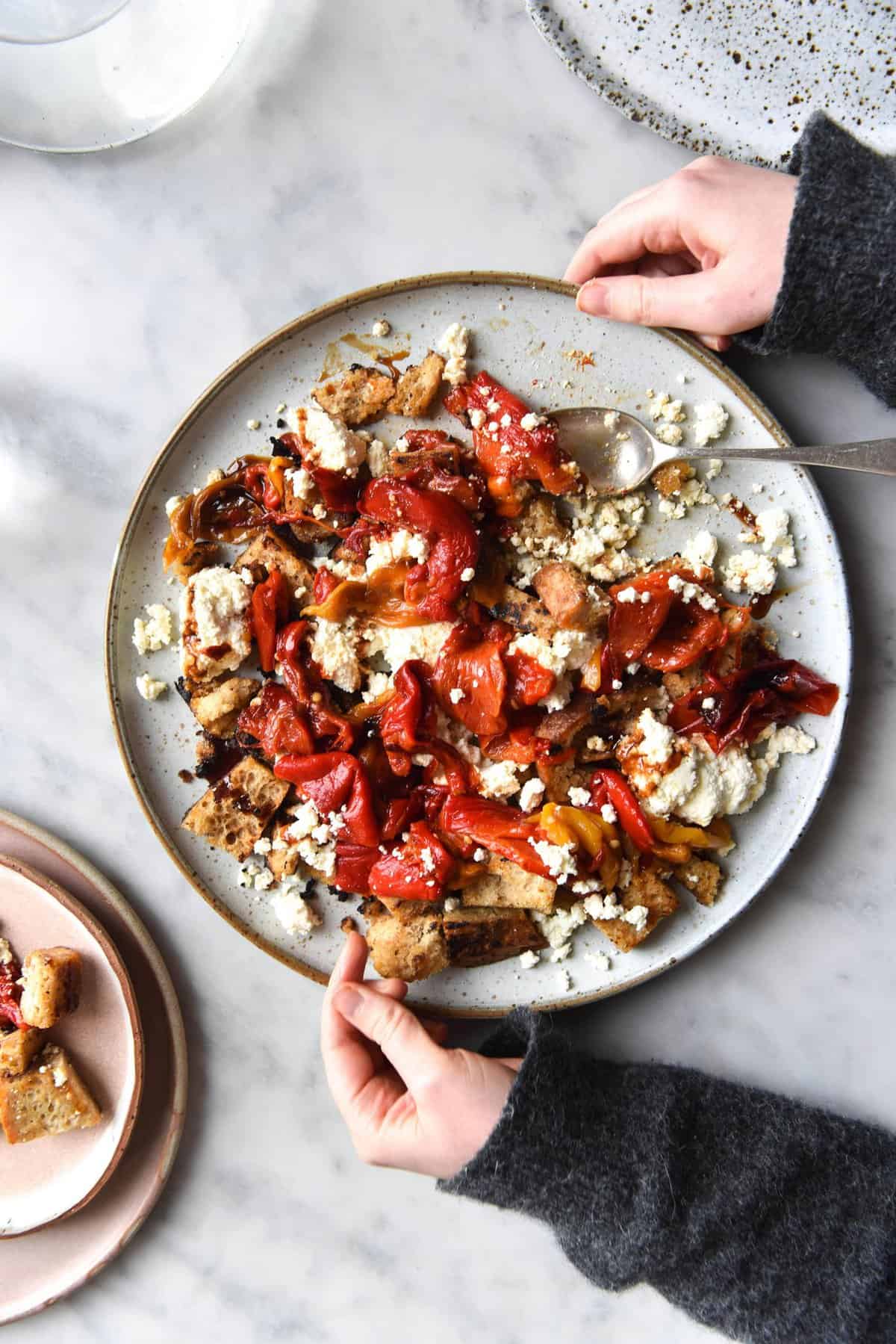 An aerial image of a plate of gluten free sourdough croutons with homemade lactose free ricotta, roasted capsicums and a balsamic glaze. The plate sits atop a white marble table and hands extend out to hold the plate. It surrounded by water glasses and extra plates.