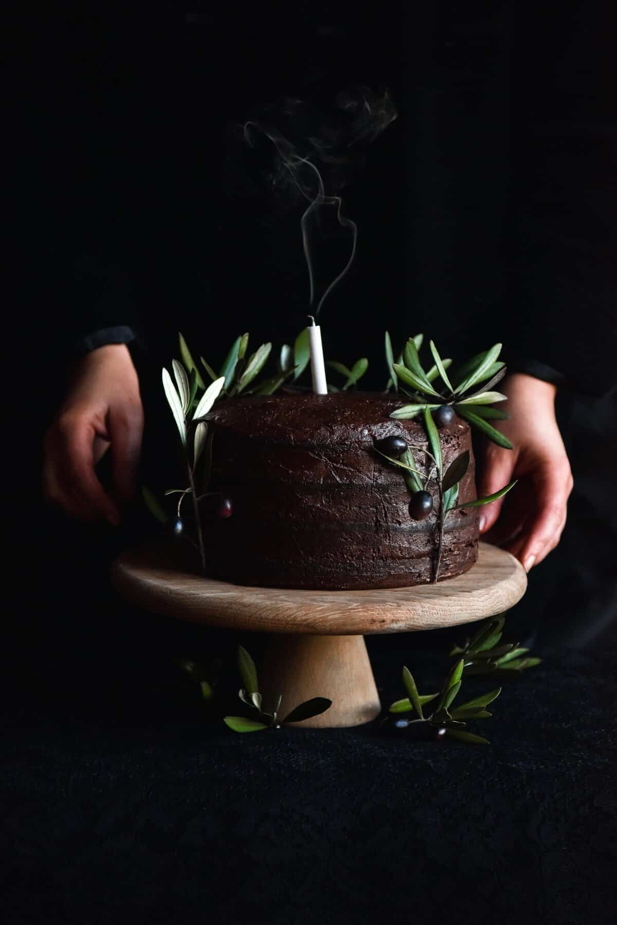 A side on moody image of a gluten free chocolate cake decorated with olive leaves on a wooden stand. A person stands behind the cake and holds the cake stand on both sides. 
