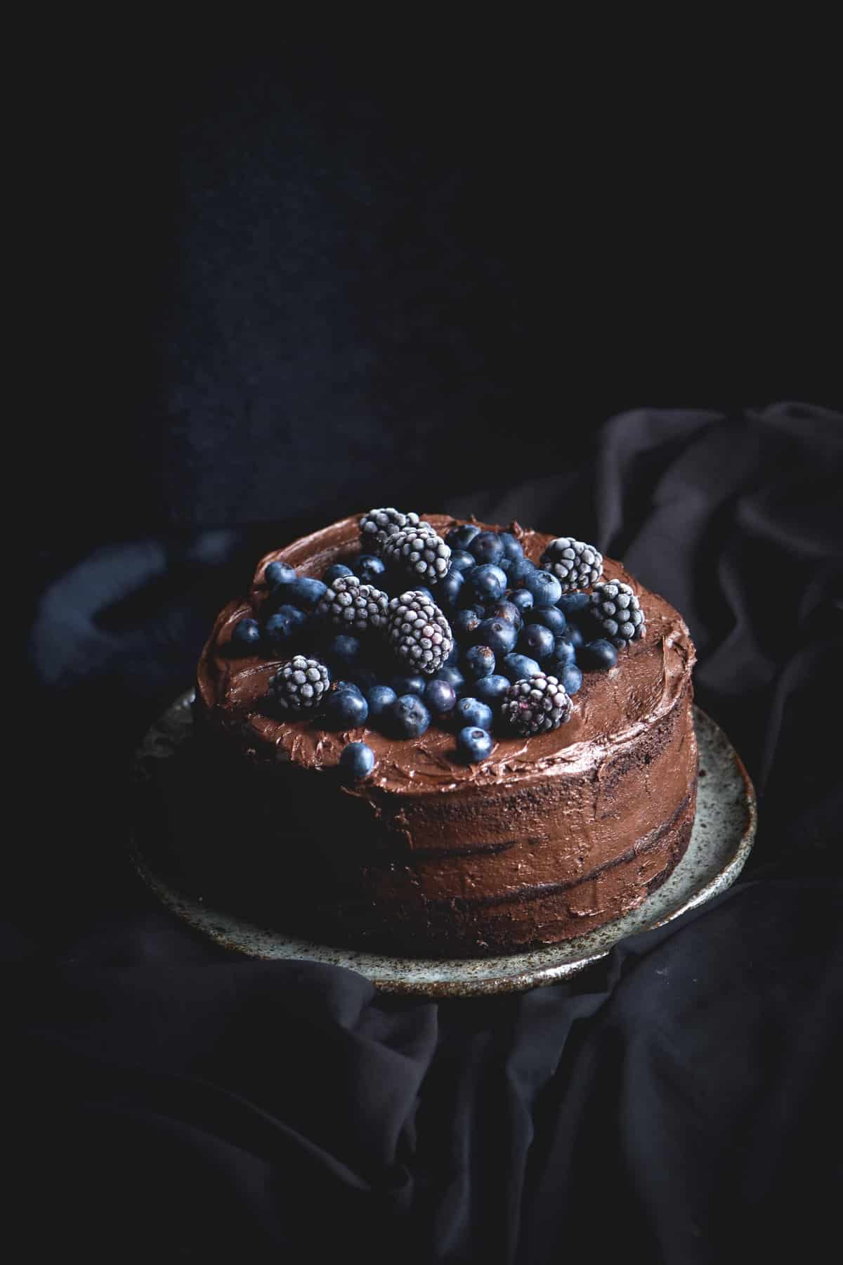 A gluten free chocolate layer cake topped with blueberries and blackberries, set against a black backdrop