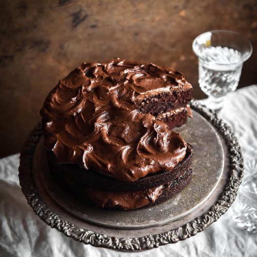 A side on image of a gluten free chocolate cake on a silver cake stand atop a white linen tablecloth. The cake has a slice taken out revealing the soft crumb and waves of chocolate buttercream. It stands against a rusty deep red backdrop