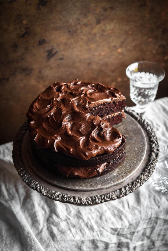 A side on image of a gluten free chocolate cake on a silver cake stand atop a white linen tablecloth. The cake has a slice taken out revealing the soft crumb and waves of chocolate buttercream. It stands against a rusty deep red backdrop