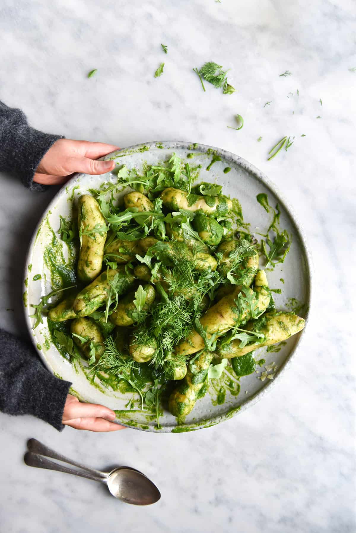 An aerial view of a super Easy Green Potato Salad on a large white serving plate atop a white marble table. Two hands in a grey jumper extend from the left side of the image to hold the plate