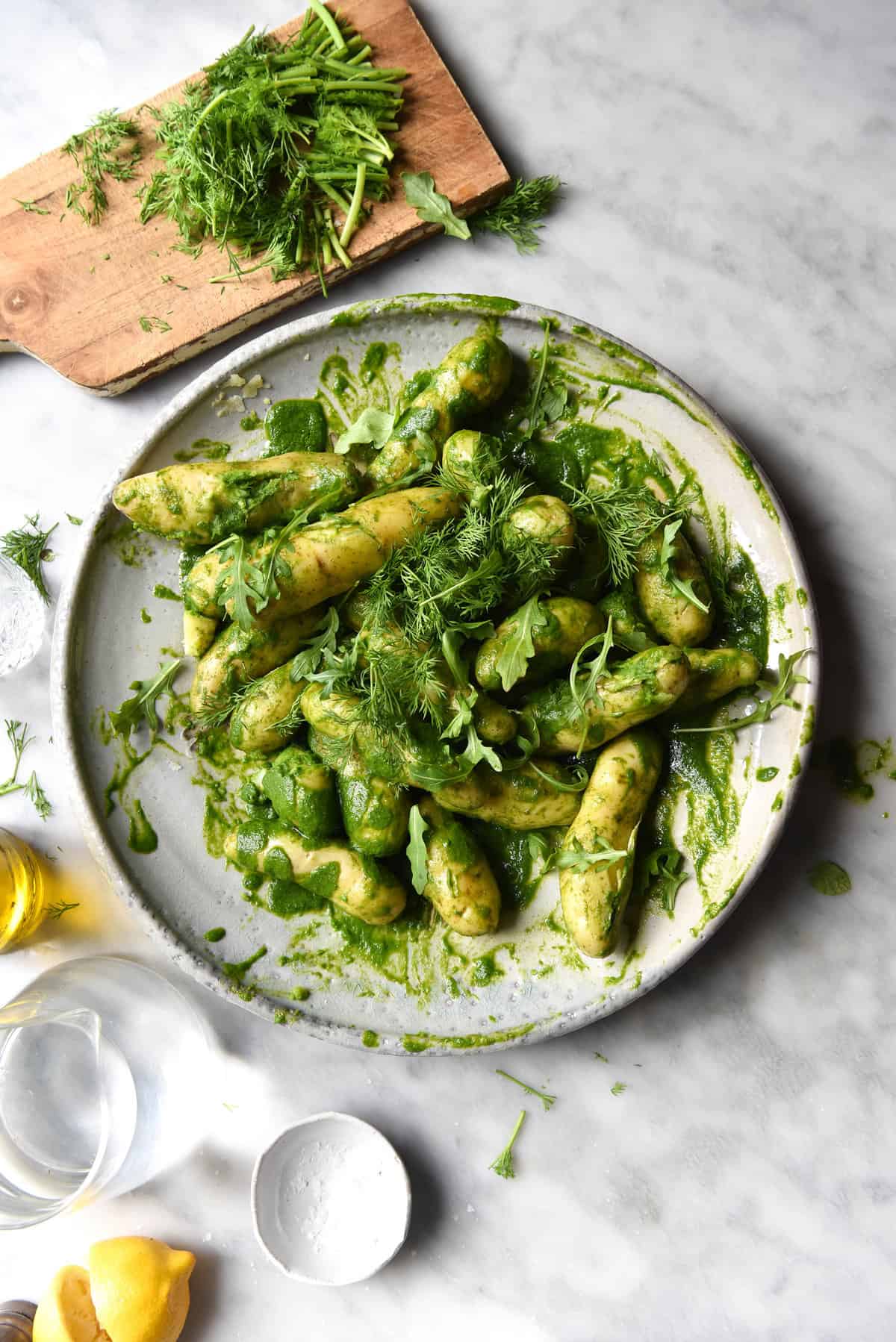 An aerial image of a green potato salad on a large white ceramic plate atop a white marble table. The table is casually strewn with herbs, lemon and glasses of water, and a herb topped chopping board sits to the top left of the salad. 