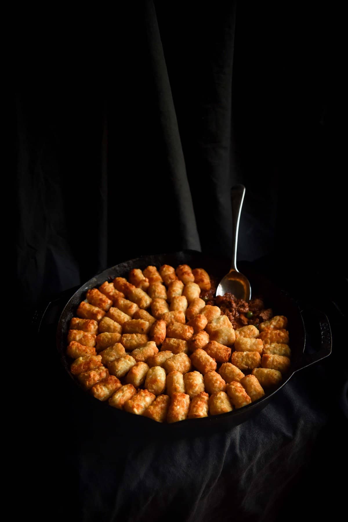 A dark and moody shot of a vegan Shepherd's pie in a black skillet topped with tater tots. 