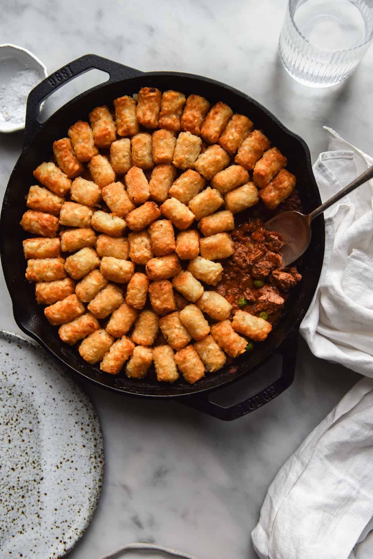 A vegan Shepherd's pie in a skillet topped with tater tots. The skillet sits on a white marble table surrounded by white plates and linen napkins
