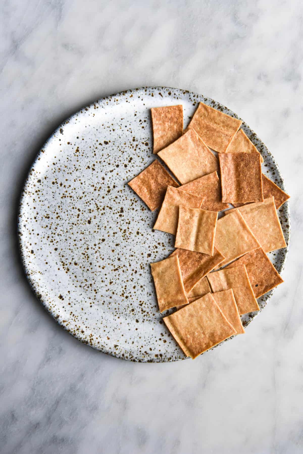 An aerial image of gluten free sourdough starter discard crackers on a white speckled ceramic plate atop a white marble table.