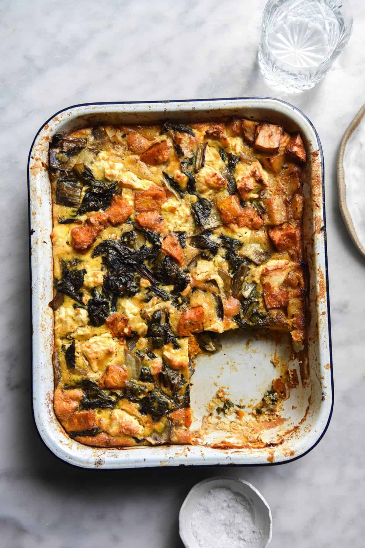 An aerial view of a FODMAP friendly lemony kale, feta, roasted pumpkin and potato frittata in a white rectangular baking dish on a white marble table. 