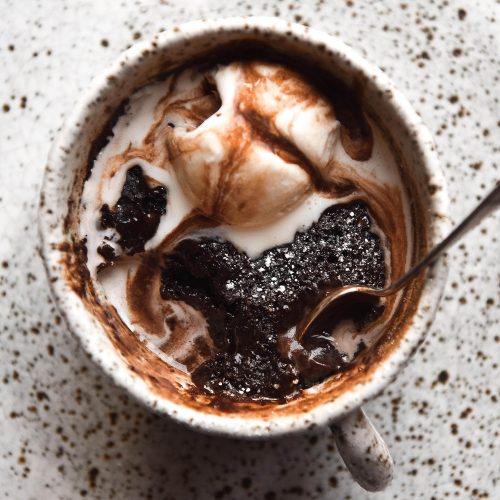 An aerial view of a gluten free chocolate mug cake in a white speckled mug atop a white speckled plate. The mug cake is topped with melting vanilla ice cream and chocolate sauce.