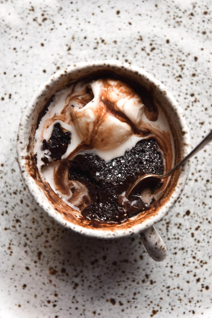 An aerial view of a gluten free chocolate mug cake in a white speckled mug atop a white speckled plate. The mug cake is topped with melting vanilla ice cream and chocolate sauce.