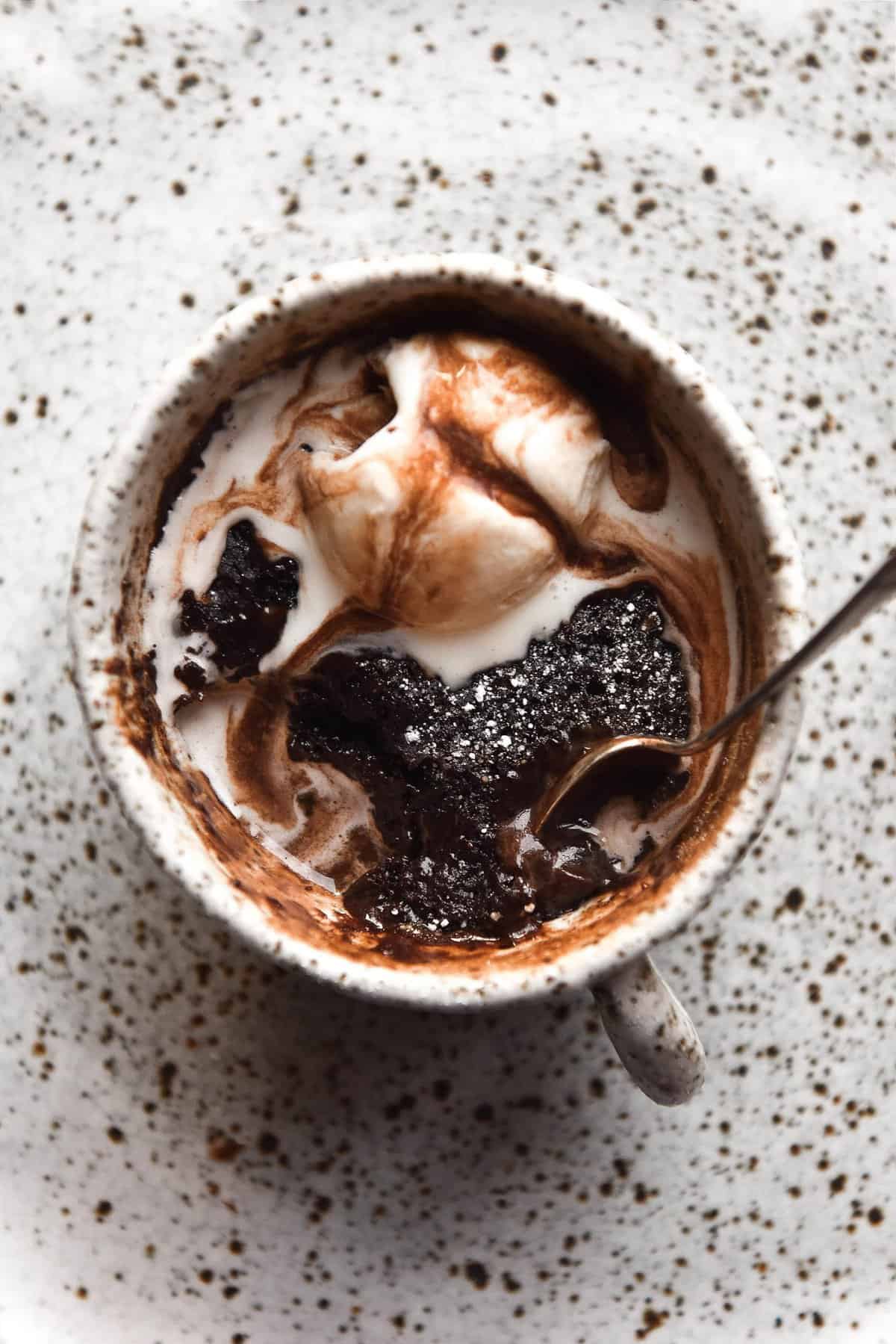 An aerial view of a gluten free chocolate mug cake in a white speckled mug atop a white speckled plate. The mug cake is topped with melting vanilla ice cream and chocolate sauce. 