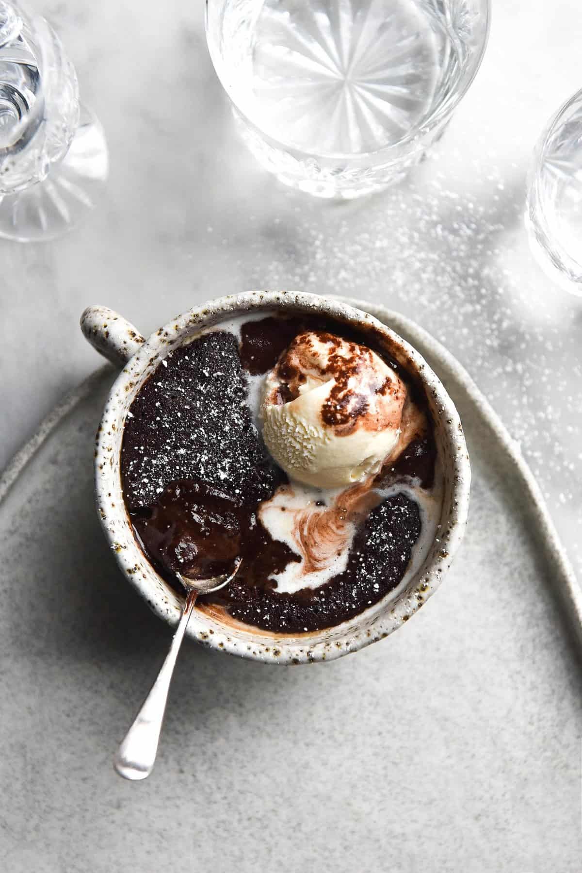 An aerial image of a refined sugar free and gluten free vegan chocolate mug cake in a white speckled ceramic mug atop a white ceramic plate. The mug cake is topped with vanilla ice cream which has melted and swirled into the chocolate mug cake. 