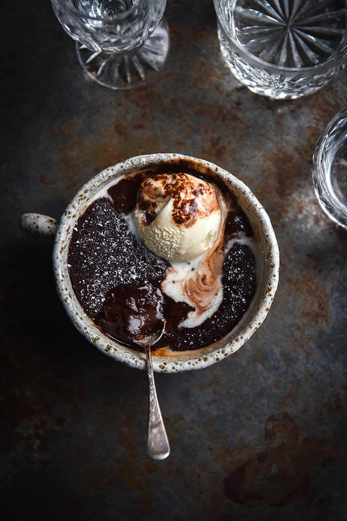 A moody image of a chocolate mug cake on a moody steel backdrop. The mug cake is topped with melting vanilla ice cream.