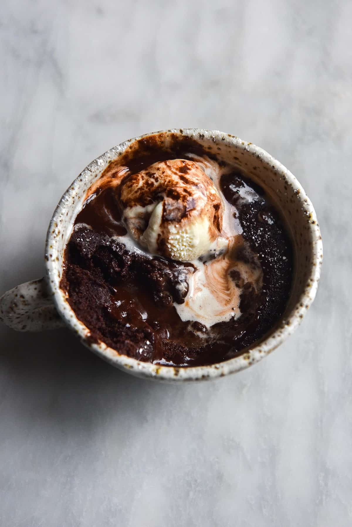 An aerial image of a chocolate mug cake topped with melting vanilla ice cream on a white marble table.
