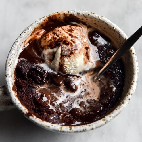 An aerial view of a gluten free low FODMAP chocolate mug cake in a white speckled ceramic mug atop a white marble table. The mug cake is topped with vanilla ice cream and chocolate sauce, which have melted together to form swirling pools on top of the cake. A spoon sticks into the cake from the right side of the image.