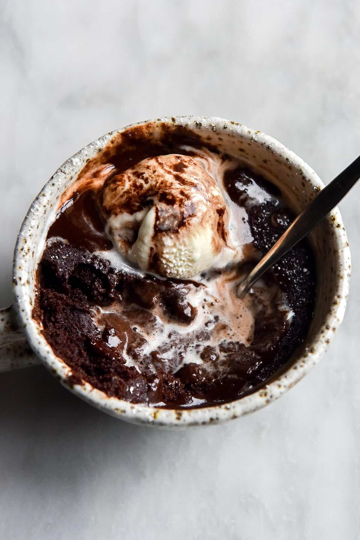 An aerial view of a gluten free low FODMAP chocolate mug cake in a white speckled ceramic mug atop a white marble table. The mug cake is topped with vanilla ice cream and chocolate sauce, which have melted together to form swirling pools on top of the cake. A spoon sticks into the cake from the right side of the image.