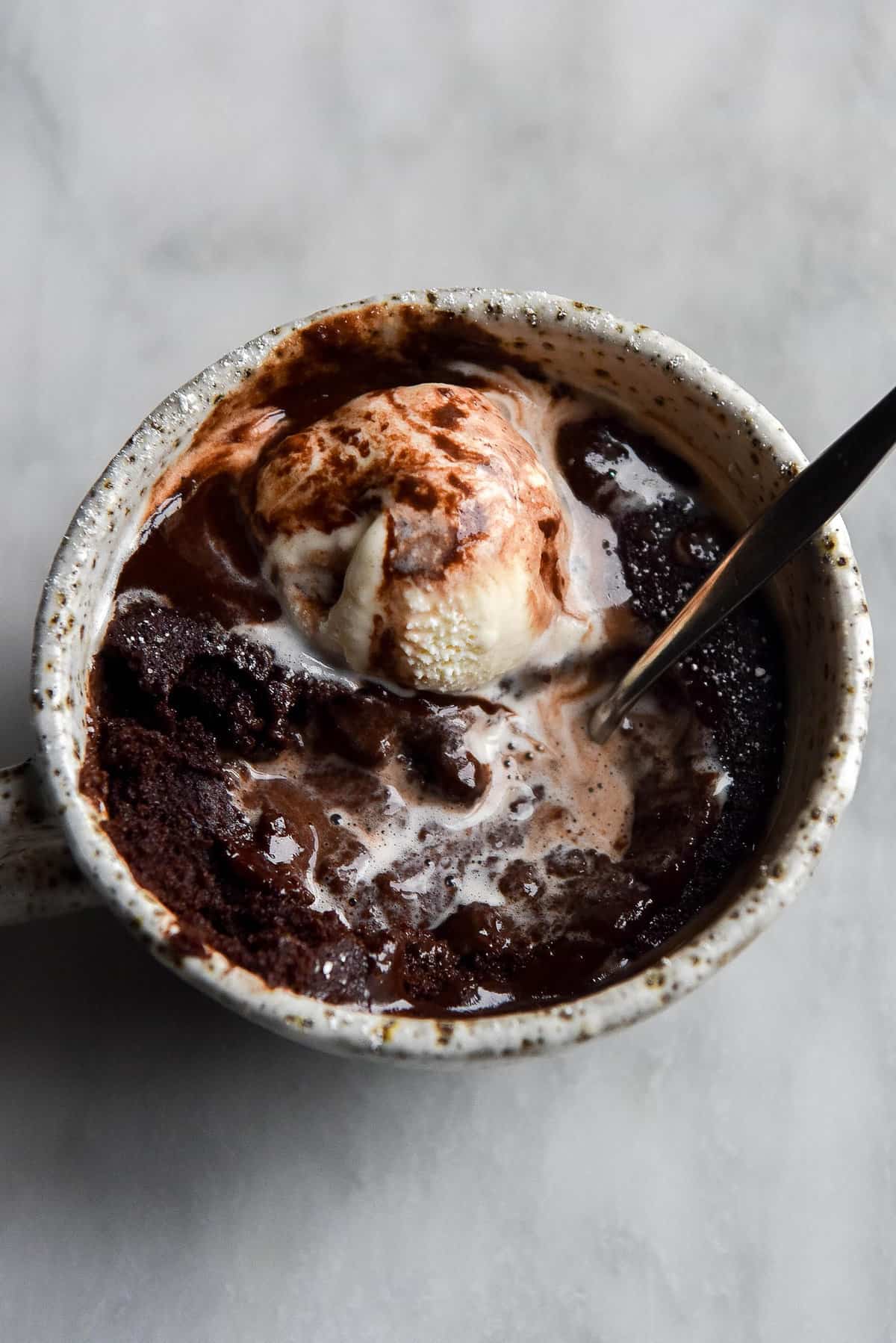 A close up aerial image of a gluten free vegan chocolate mug cake in a white speckled ceramic mug atop a white marble table. The mug cake is topped with melting vanilla ice cream and a sprinkling of icing sugar. 